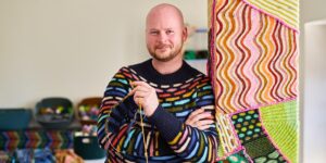 Stephen West in one of his sweaters, holding knitting, surrounded by shawls